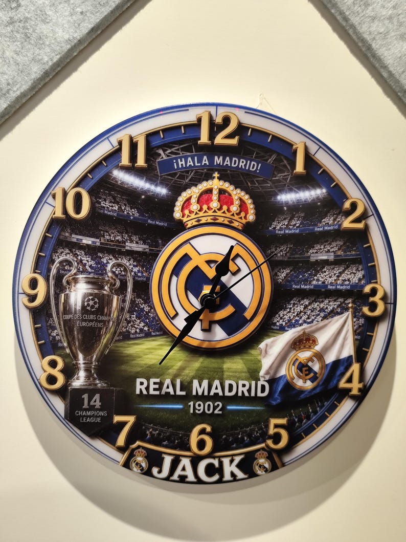 May include: A round wall clock featuring a Real Madrid soccer stadium background, the team logo with a crown, a Champions League trophy, and a flag. The clock face displays the text &iexcl;HALA MADRID!, REAL MADRID 1902, 14 CHAMPIONS LEAGUE, and JACK.