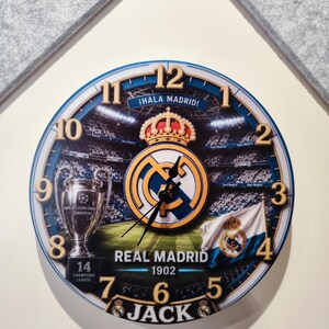 May include: A round wall clock featuring a Real Madrid theme with a stadium background, the team crest, a trophy, and a flag. The clock face displays the text &iexcl;HALA MADRID!, REAL MADRID 1902, 14 CHAMPIONS LEAGUE, and the name JACK in bold letters.