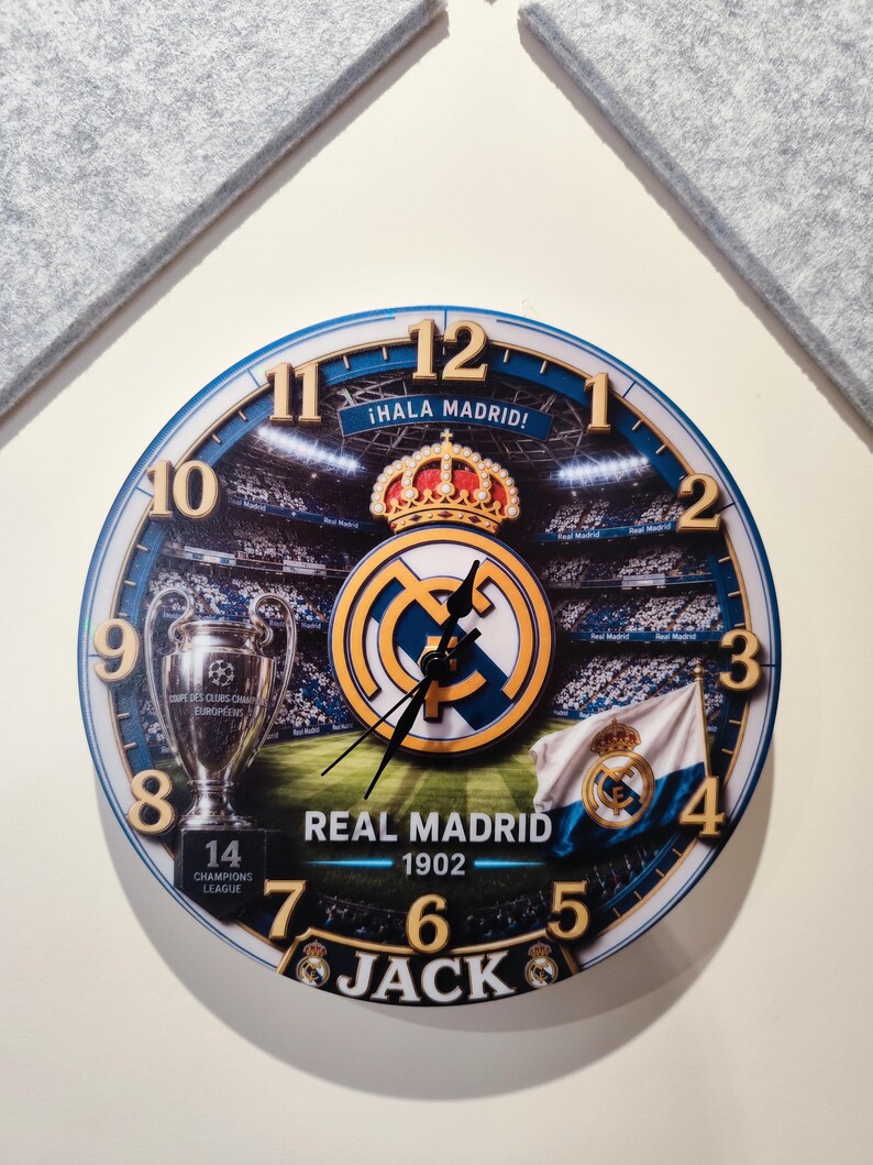 May include: A round wall clock featuring a Real Madrid theme with a stadium background, the team crest, a trophy, and a flag. The clock face displays the text &iexcl;HALA MADRID!, REAL MADRID 1902, 14 CHAMPIONS LEAGUE, and the name JACK in bold letters.
