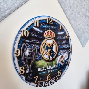 May include: A round wall clock featuring a Real Madrid theme with a stadium background, the team logo, a Champions League trophy, and the text "&iexcl;HALA MADRID!", "REAL MADRID 1902", and "JACK". The clock has gold-colored numbers and black hands.