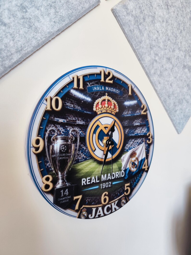 May include: A round wall clock featuring a Real Madrid theme with a stadium background, the team logo, a Champions League trophy, and the text "&iexcl;HALA MADRID!", "REAL MADRID 1902", and "JACK". The clock has gold-colored numbers and black hands.