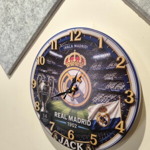 May include: A round wall clock featuring a Real Madrid soccer stadium graphic, the team logo, a crown, a trophy, and a flag. The clock face displays the text "&iexcl;HALA MADRID!", "REAL MADRID", "1902", and "JACK" with gold-colored numbers from 1 to 12.