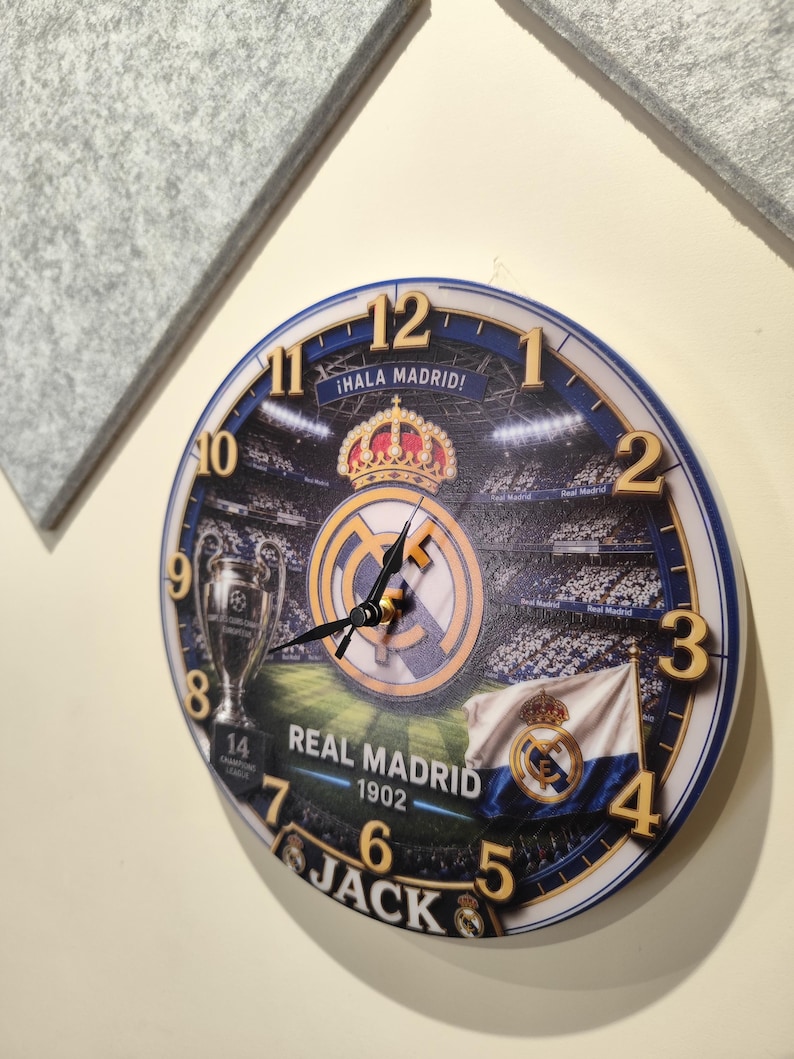 May include: A round wall clock featuring a Real Madrid soccer stadium graphic, the team logo, a crown, a trophy, and a flag. The clock face displays the text "&iexcl;HALA MADRID!", "REAL MADRID", "1902", and "JACK" with gold-colored numbers from 1 to 12.