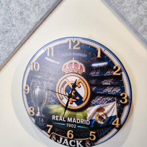 May include: A round wall clock featuring a Real Madrid theme with the club crest, a stadium background, the Champions League trophy, and a flag. The clock face displays the text Hala Madrid, Real Madrid 1902, and the name Jack with gold numbers.