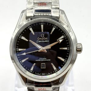 Könnte beinhalten: Eine silberfarbene Armbanduhr aus Edelstahl mit dunkelblauem Zifferblatt, Omega Seamaster-Logo, Co-Axial Master Chronometer-Schriftzug, Datumsanzeige auf der Sechs-Uhr-Position und einem in Schutzfolie eingewickelten Metallgliederarmband.