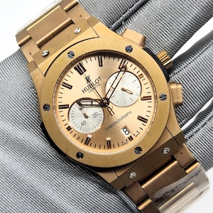 Könnte beinhalten: Eine goldfarbene Hublot Geneve Chronographenuhr mit metallischem Finish, ausgestattet mit einem runden Zifferblatt mit Hilfszifferblättern, Stabindizes und einer Datumsanzeige auf der Vier-Uhr-Position. Die Uhr hat ein passendes Metallgliederarmband.