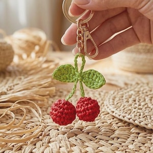 May include: A crochet cherry keychain featuring two small, textured red spheres attached to green crocheted leaves and stems. The fruit is connected to a gold-toned metal split ring and a lobster clasp, providing a secure attachment for keys or bags.