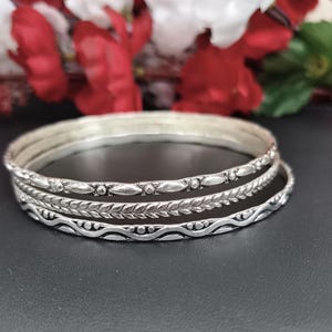 May include: A set of three silver-toned metal bangles featuring distinct textures. The top bangle has a repeating leaf-like pattern, the middle one displays a braided design, and the bottom bangle showcases a wavy line with small dot accents.