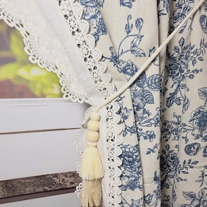 Könnte beinhalten: Cremefarbener Vorhang mit blauem Blumenmuster und weißer Spitzenborte. Eine dekorative Quaste mit Holzperlen und mehrschichtigen beigen Quasten hängt an der Gardinenbindung. Der Vorhang ist aus einem leinenartigen Stoff gefertigt.
