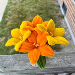 May include: A bouquet of three handcrafted flowers made from fuzzy pipe cleaners. Two flowers are bright yellow and one is vibrant orange. Each flower features six petals and a central cluster of small yellow stamens. A single green leaf is visible.