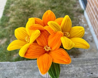 Puede incluir: Un ramo de tres flores artesanales hechas con limpiapipas difusos. Dos flores son de color amarillo brillante y una es de color naranja vibrante. Cada flor tiene seis pétalos y un centro con pequeños estambres amarillos. Se ve una hoja verde.