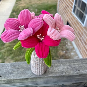 Op de afbeelding: Een boeket van handgemaakte pijpenragerbloemen in tinten felroze, middenroze en lichtroze, met groene pijpenragerbladeren en witte meeldraden, geschikt in een kleine, getextureerde, gebroken witte gehaakte vaas met een afgeronde onderkant.
