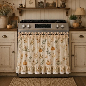 May include: A cream-colored stove curtain featuring a repeating pattern of orange pumpkins, green leaves, and wheat stalks. The curtain is adorned with a row of tan and white striped bows along the top edge and a white lace trim at the bottom hem.