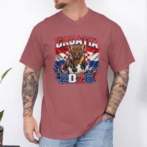 May include: A dusty rose V-neck t-shirt featuring a graphic of a snarling pine marten with a soccer ball, the text CROATIA, and the year 2026. The design incorporates the red and white checkered pattern of the Croatian flag with blue accents.