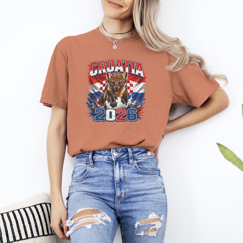 May include: A terracotta-colored short-sleeve t-shirt featuring a graphic print of a snarling animal with a soccer ball, the text CROATIA, the year 2026, and a red, white, and blue checkered flag background with blue and red paint splatter accents.