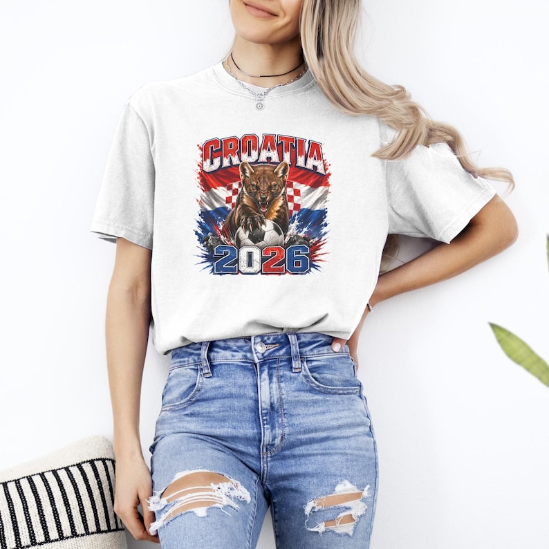 May include: A white short-sleeved t-shirt featuring a graphic print of a snarling marten over a Croatian flag background, a soccer ball, and the text CROATIA 2026 in red, white, and blue. The shirt is paired with light blue distressed denim jeans.