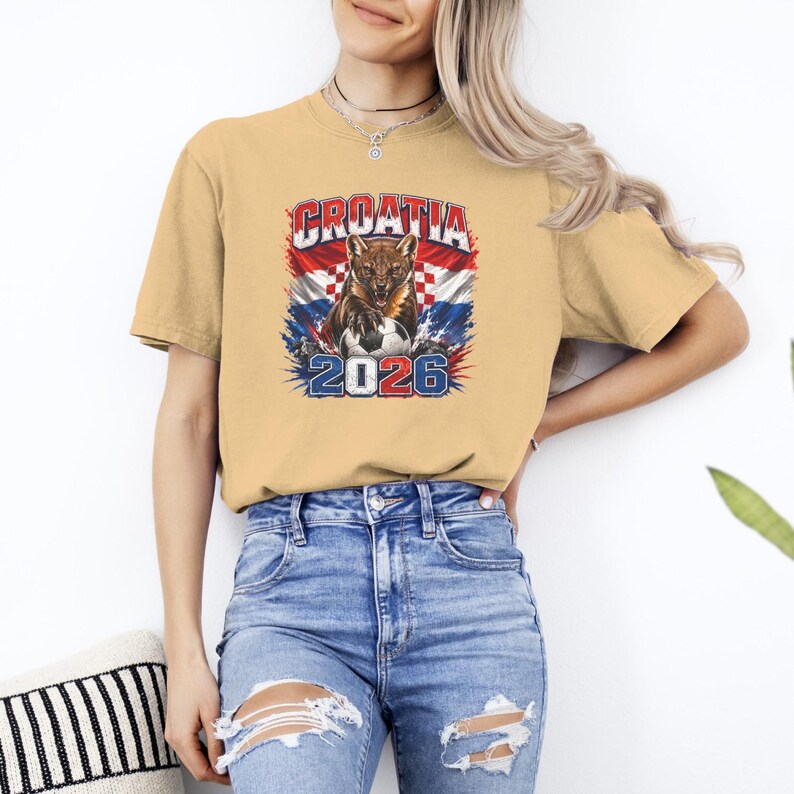 May include: A tan short-sleeve t-shirt featuring a graphic print of a snarling animal with a soccer ball. The design includes the text CROATIA and 2026, set against a background of the Croatian flag with red, white, and blue colors.
