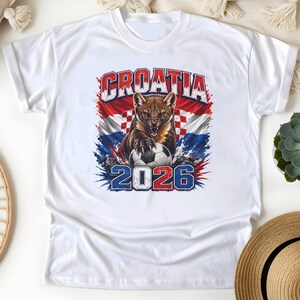 May include: A white short-sleeve t-shirt featuring a graphic print of a snarling pine marten with its paws on a soccer ball. The background includes the Croatian flag and the text CROATIA 2026 in bold, distressed blue and red lettering.