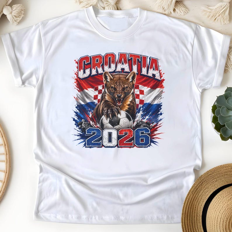 May include: A white short-sleeve t-shirt featuring a graphic print of a snarling pine marten with its paws on a soccer ball. The background includes the Croatian flag and the text CROATIA 2026 in bold, distressed blue and red lettering.