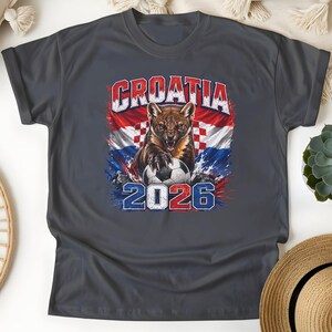 May include: A dark gray t-shirt featuring a graphic of a pine marten with a soccer ball, set against the Croatian flag. The text reads CROATIA at the top and 2026 at the bottom, with a distressed, paint-splatter effect in red, white, and blue.