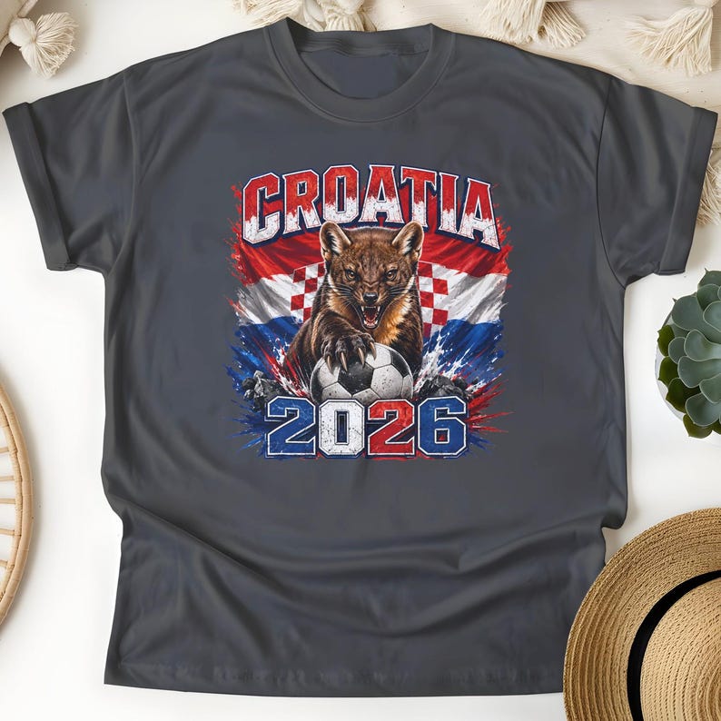 May include: A dark gray t-shirt featuring a graphic of a pine marten with a soccer ball, set against the Croatian flag. The text reads CROATIA at the top and 2026 at the bottom, with a distressed, paint-splatter effect in red, white, and blue.