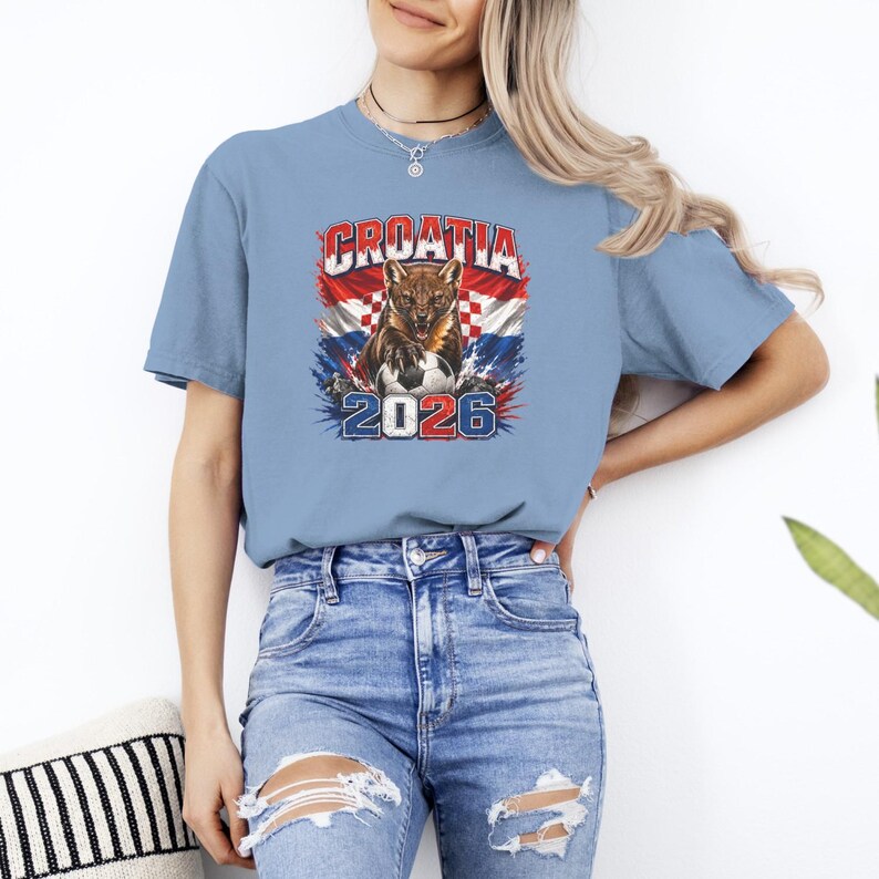 May include: A light blue short-sleeve t-shirt featuring a graphic print of a snarling animal with a soccer ball. The design includes the text "CROATIA" above the animal and "2026" below, set against a background of the Croatian flag colors.