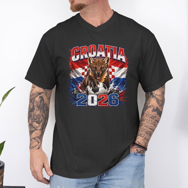 May include: A black short-sleeve t-shirt featuring a graphic of a marten animal with a soccer ball, the text CROATIA, and the year 2026. The design includes a red and white checkered pattern background with blue accents and a distressed texture.