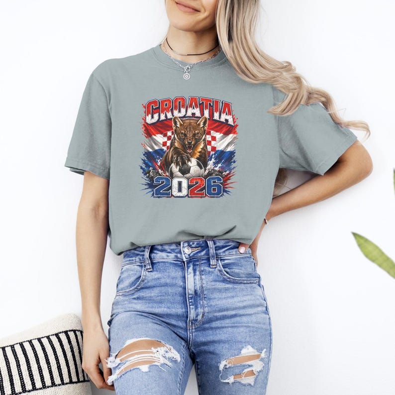 May include: A light gray short-sleeve t-shirt featuring a graphic print of a snarling lynx with a soccer ball, the text CROATIA, and the year 2026. The design incorporates the red and white checkered pattern of the Croatian flag in the background.