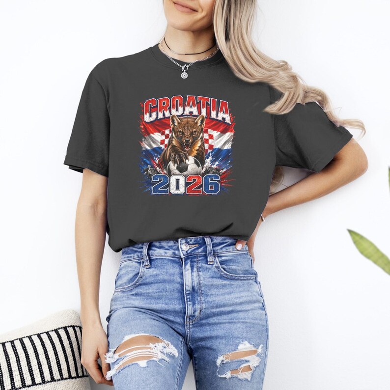 May include: A dark gray short-sleeve t-shirt featuring a graphic print of a snarling animal over a red, white, and blue checkered flag background. The text CROATIA is printed above the animal, and the number 2026 is printed below a soccer ball.