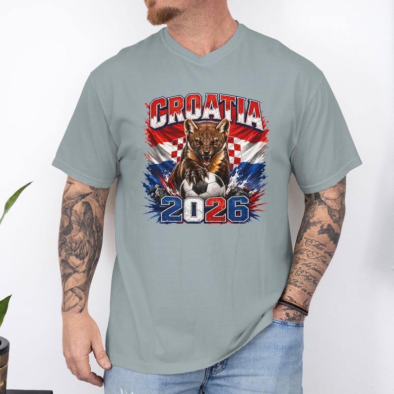 May include: A light gray short-sleeve t-shirt featuring a graphic print of a snarling pine marten with its paw on a soccer ball, set against the Croatian flag. The text CROATIA is printed above, and the year 2026 is printed below in bold blue and red.