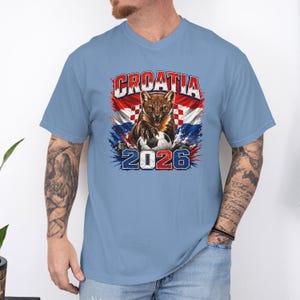 May include: A light blue V-neck t-shirt featuring a graphic of a pine marten with a soccer ball, the text CROATIA, and the year 2026. The design incorporates the red, white, and blue colors of the Croatian flag with a distressed, artistic style.
