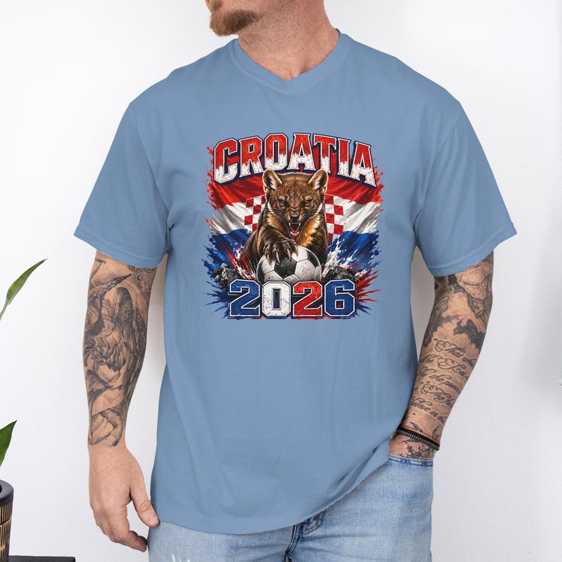 May include: A light blue V-neck t-shirt featuring a graphic of a pine marten with a soccer ball, the text CROATIA, and the year 2026. The design incorporates the red, white, and blue colors of the Croatian flag with a distressed, artistic style.