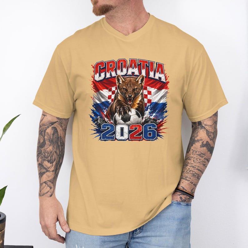 May include: A tan short-sleeve t-shirt featuring a graphic of a marten animal with a soccer ball, the text CROATIA, and the year 2026. The design includes a red and white checkered pattern background with blue accents and a distressed artistic style.