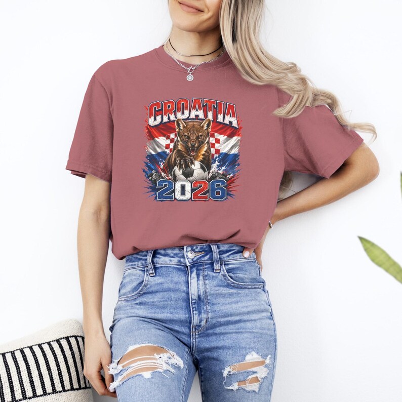 May include: A dusty rose short-sleeve t-shirt featuring a graphic of a snarling marten over a Croatian flag background with a soccer ball and the text CROATIA 2026. The shirt is paired with blue denim jeans that have distressed knee cutouts.