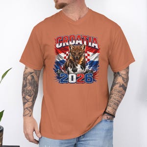 May include: A burnt orange short-sleeve t-shirt featuring a graphic print of a snarling pine marten with a soccer ball, the text CROATIA, and the year 2026. The design incorporates the red and white checkered pattern of the Croatian flag.