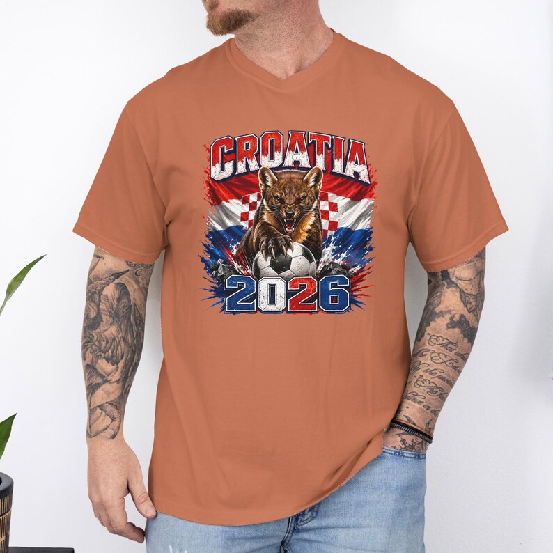 May include: A burnt orange short-sleeve t-shirt featuring a graphic print of a snarling pine marten with a soccer ball, the text CROATIA, and the year 2026. The design incorporates the red and white checkered pattern of the Croatian flag.