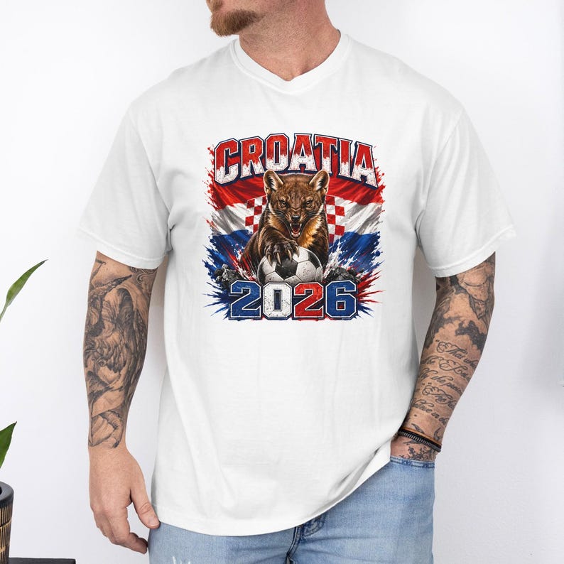 May include: A white short-sleeve t-shirt featuring a graphic print of a snarling marten with a soccer ball. The design includes the text CROATIA and 2026, set against a background of the Croatian flag with red, white, and blue paint splatters.