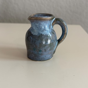 May include: A small ceramic creamer pitcher featuring a rounded body and a curved handle. The glaze displays a blend of deep blue and earthy brown tones, with a lighter, mottled blue finish around the rim and upper section of the vessel.