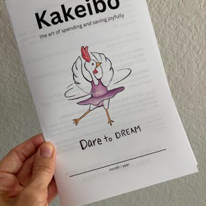 May include: A white booklet titled Kakeibo the art of spending and saving joyfully. The cover features a cartoon illustration of a white chicken wearing a purple tutu and dancing. Below the chicken, the text reads Dare to DREAM and month / year.