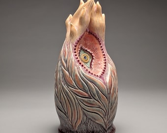 May include: A ceramic vase with a unique design. The vase features a central eye-like opening with a detailed iris and a surrounding border of small red beads. The exterior is adorned with leaf-like patterns in shades of gray, peach, and brown.