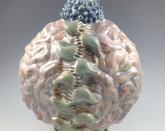 May include: A spherical ceramic vessel with a textured, brain-like surface in shades of pink and tan. A vertical row of green, leaf-shaped protrusions runs down the center. The top features a dark blue, spiky collar and a white, twisted finial.