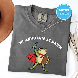 May include: A charcoal gray t-shirt featuring a graphic of a green frog wearing a red cape, holding a blue marker and a brown clipboard. The text "WE ANNOTATE AT DAWN" is printed in white above the frog. The shirt has a crew neckline and a tag.