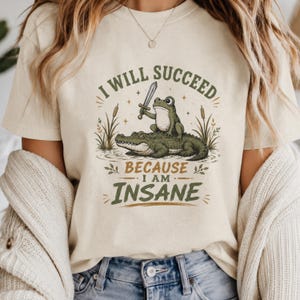 Op de afbeelding: Een crèmekleurig t-shirt met een afbeelding van een kikker die een zwaard vasthoudt terwijl hij op de rug van een alligator zit. De tekst luidt I WILL SUCCEED BECAUSE I AM INSANE. Het ontwerp bevat lisdodden en kleine sterren in groen en bruin.