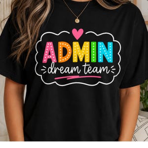 May include: A black t-shirt featuring a graphic design with a pink heart above the word ADMIN in multicoloured, polka-dotted letters. Below, the text dream team is written in white script, all enclosed within a white scalloped border.