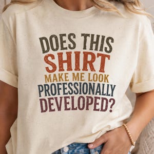 May include: A cream-colored short-sleeve t-shirt with a speckled texture. The front features distressed, multi-colored text that reads: DOES THIS SHIRT MAKE ME LOOK PROFESSIONALLY DEVELOPED? The lettering uses dark green, orange, gold, blue, and maroon.