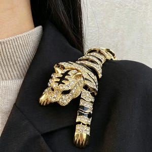 May include: A gold-toned tiger brooch featuring clear crystal rhinestones and black enamel stripes. The articulated design depicts a tiger in a prowling position with its head turned, showcasing detailed metalwork and sparkling stone accents.