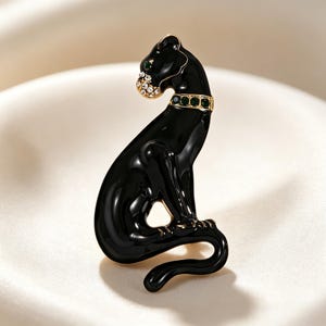 May include: A black panther brooch featuring a glossy enamel finish, a gold-tone collar set with green crystals, and a muzzle accented with clear white rhinestones. The feline figure is depicted in a seated position with its tail curved downward.