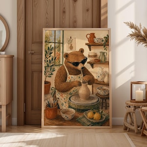 Op de afbeelding: Een ingelijste kunstdruk toont een bruine beer met een zonnebril en een schort, die potten bakt op een draaischijf. Het kunstwerk heeft een neutraal kleurenpalet met aardewerk, planten en citroenen, wat een gezellig, artistiek thema creëert.