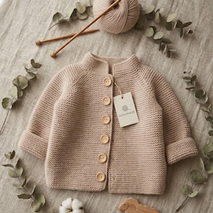 May include: A beige knit cardigan with long sleeves, a high neckline, and six light-colored wooden buttons. A rectangular tag attached to the garment features a yarn ball icon and the text HANDMADE. The cardigan is displayed with a ball of yarn, two knitting needles, a cotton boll, and a wooden sheep figurine.