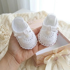 May include: A pair of white, crocheted baby shoes with a delicate floral design and pearl accents. The shoes are displayed in a decorative gift box with a satin ribbon. The overall theme is baby and infant footwear.